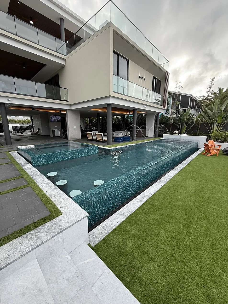 The same backyard finished: teal glass-tile infinity pool and spa, marble coping, turf and pavers