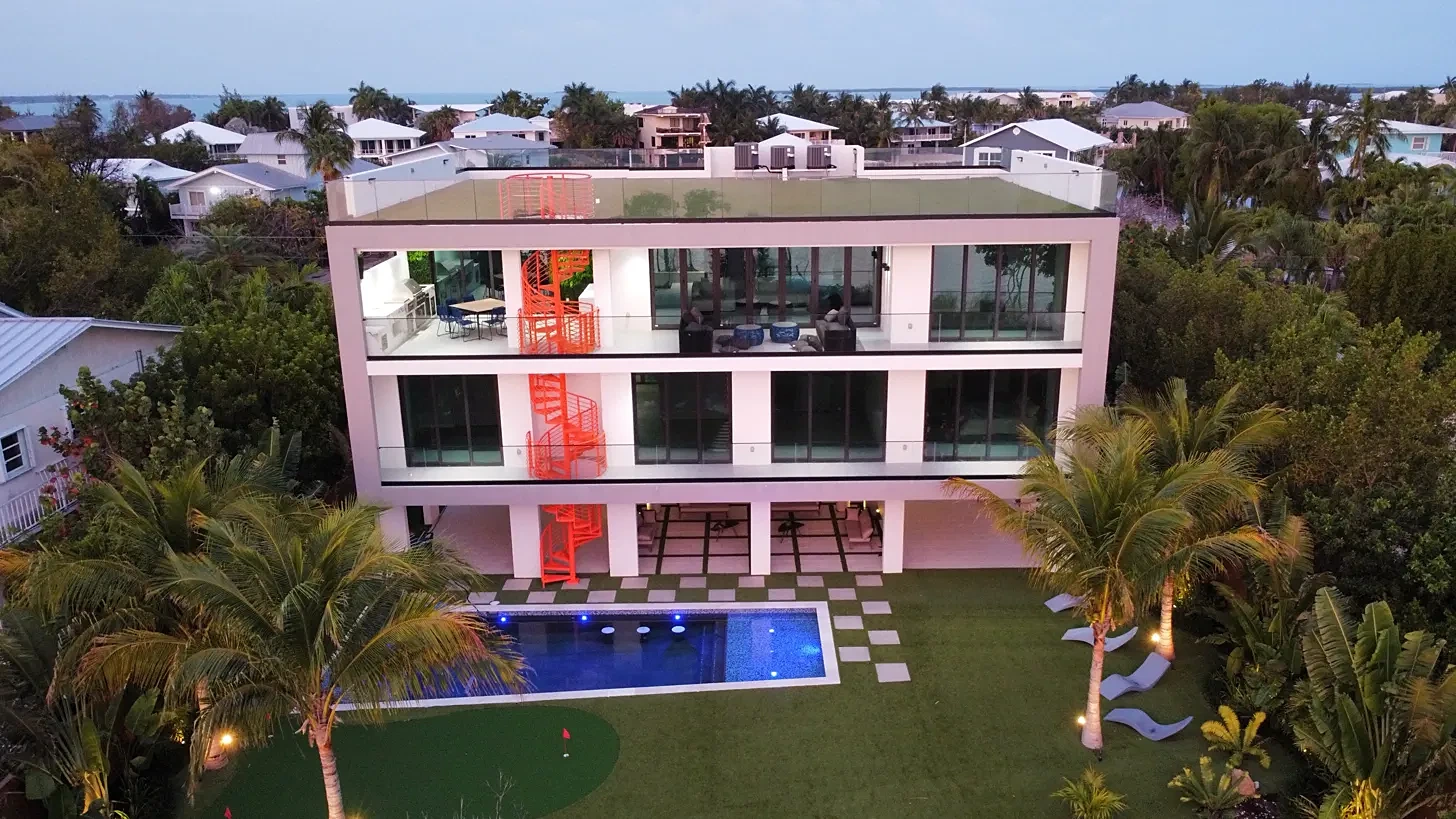 Aerial view at dusk of a modern three-story Florida Keys home with an orange spiral staircase, lit pool, putting green and palms, built by Buildia Corp
