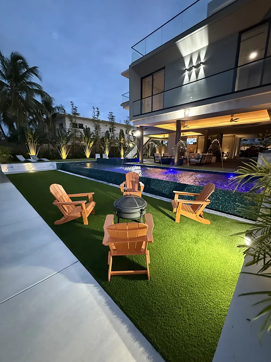 Outdoor living at night: lit infinity-edge pool, fire pit with Adirondack chairs and turf