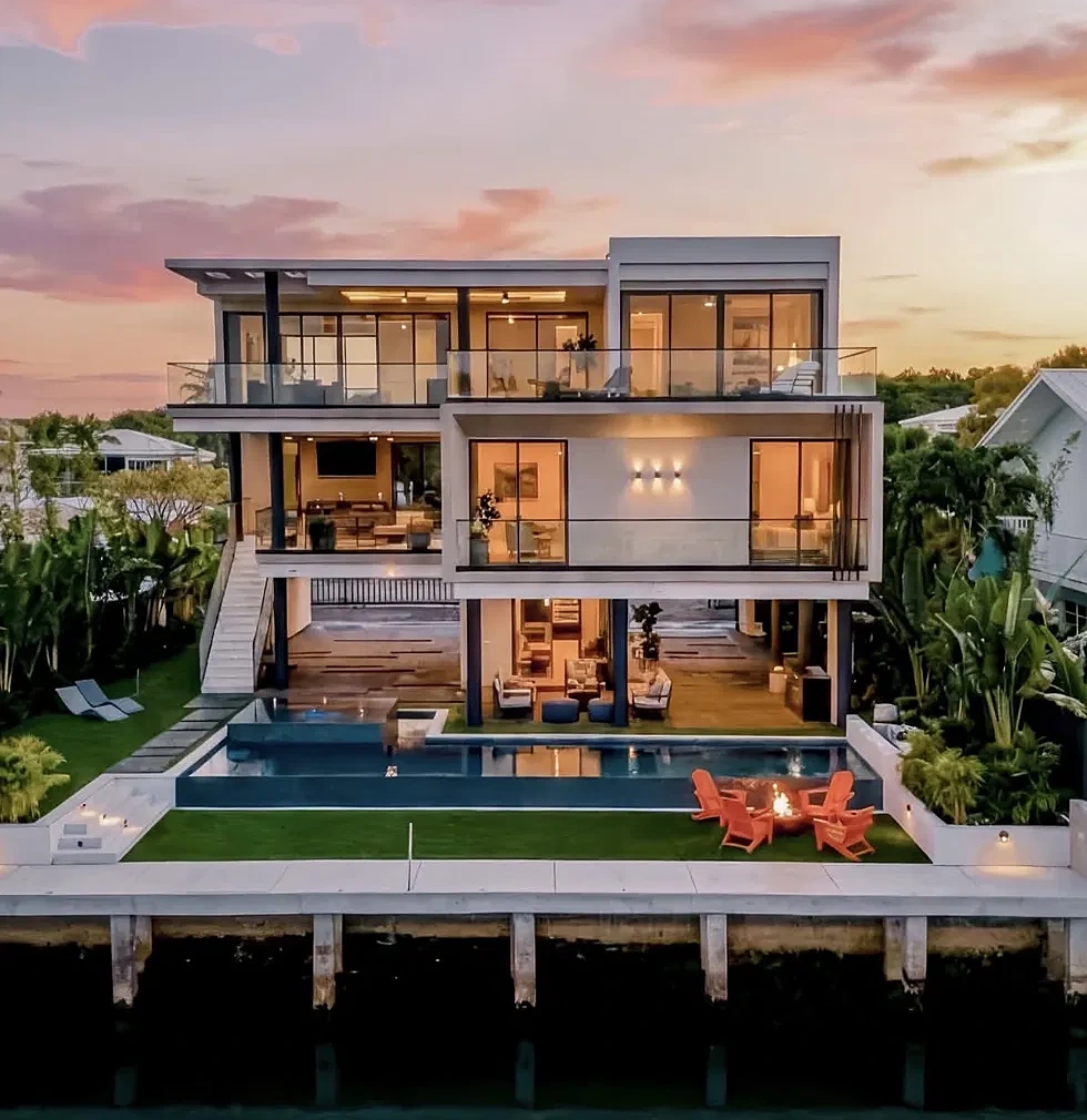 Modern waterfront home at sunset with infinity pool, fire pit and turf lawn