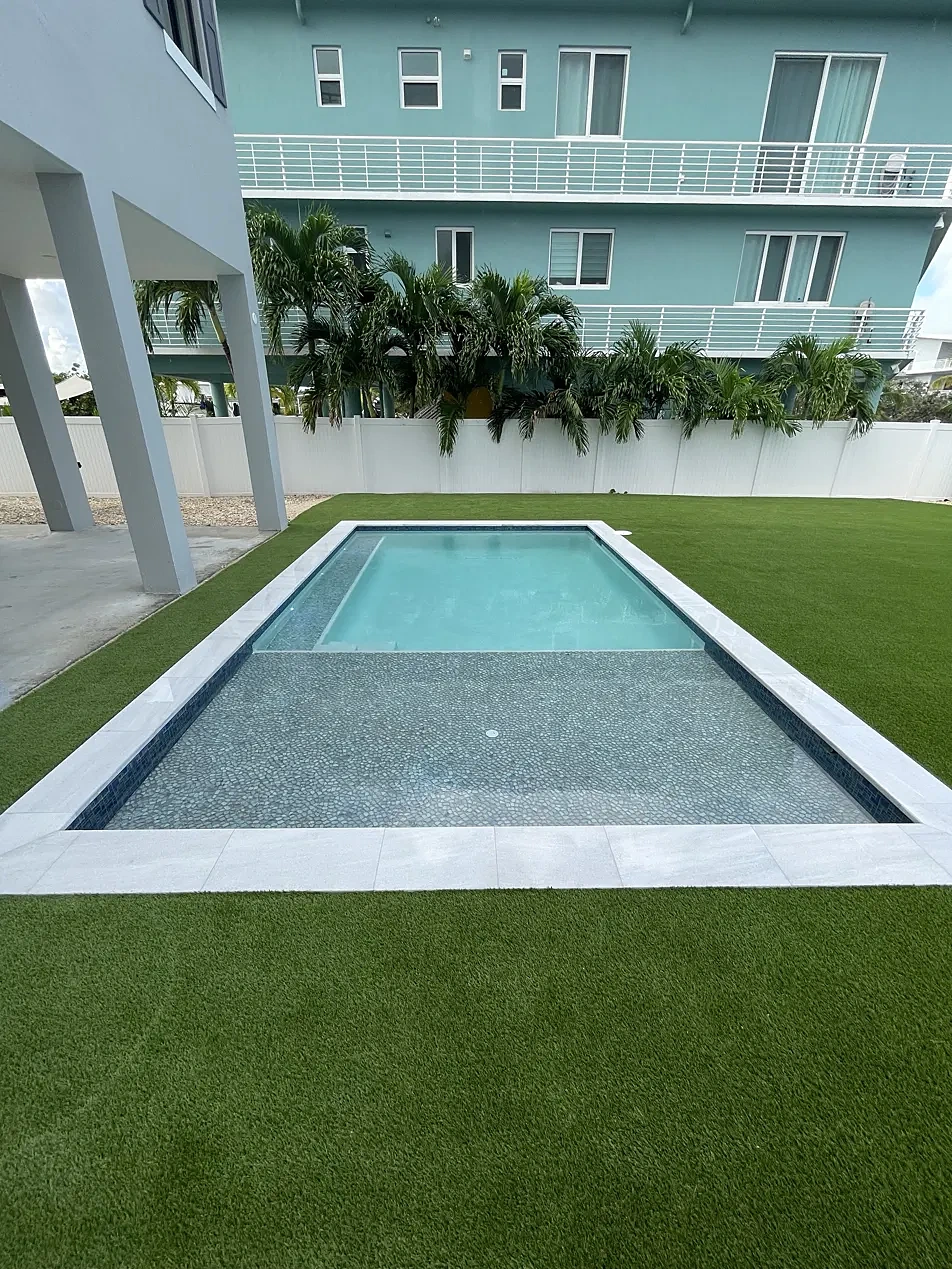Rectangular pool with sun shelf, marble coping and artificial turf beside white privacy fence